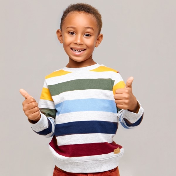 Positive human emotions reaction and feelings emotional happy dark skinned boy in multi colored jumper making thumbs up gesture expressing agreement approval giving his like smiling broadly